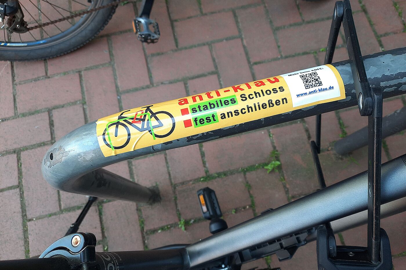 Der Aufkleber in Originalgröße mit den beiden Kern-Sicherungsmaßnahmen und dem QR-Code auf einem Fahrradbügel, gleich neben einem Faltschloss-gesicherten Fahrrad, fest angeschlossen an einem der vielen Fahrradbügel in Langenhagens Stadtgebiet
