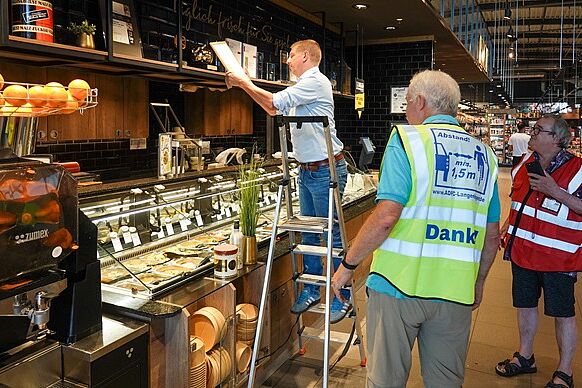 Das Goldene RadPedal Über der Salatbar, gleich links vom Eingang: EDEKA-Marktleiter André Fiolek positioniert das "Goldene RadPedal" und die Urkunde bei den anderen Auszeichnungen, unterstützt von Dr. Reinhard Spörer (gelbe ADFC-Weste) und Reinhard Wolf (rote ADFC-Weste)