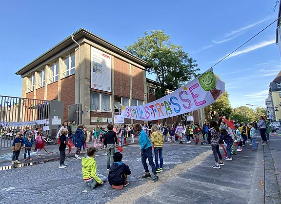 Kinder verschiedenen Alters spielen und bewegen sich auf einer gepflasterten Straße vor einem zweistöckigen Backsteingebäude. Über der Straße hängt ein buntes Banner mit der Aufschrift „Schulstraße“ und einem grünen Baum-Symbol. Im Hintergrund sind Erwachsene zu sehen, die die Aktion begleiten.