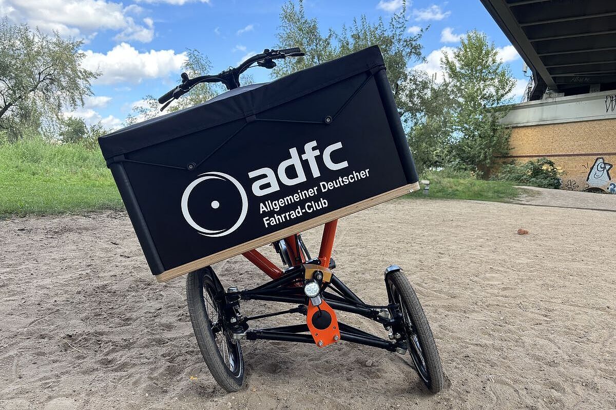 Ein dreirädriges Lastenrad mit schräg geneigter Transportbox steht auf einem sandigen Weg.