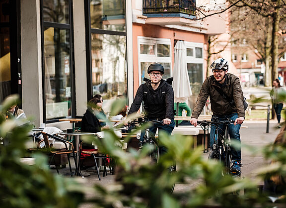 Zwei Männer mit Helm fahren auf einem Radweg im urbanen Raum